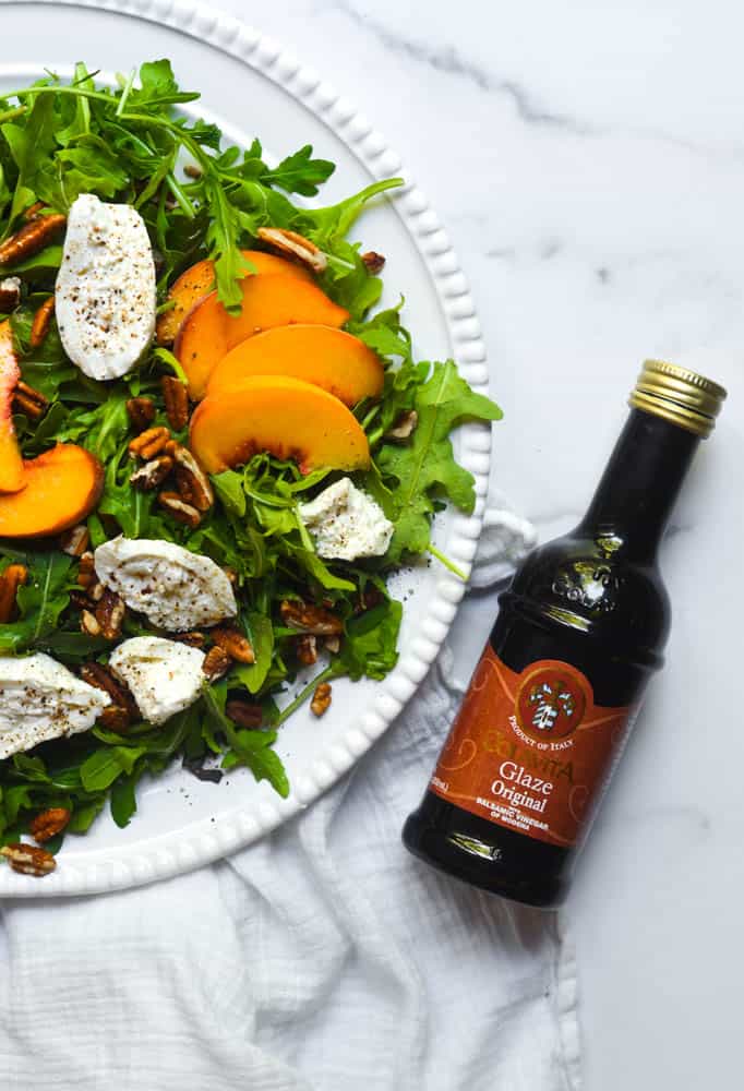 peach salad with burrata, arugula and pecans on white plate next to a bottle of balsamic glaze