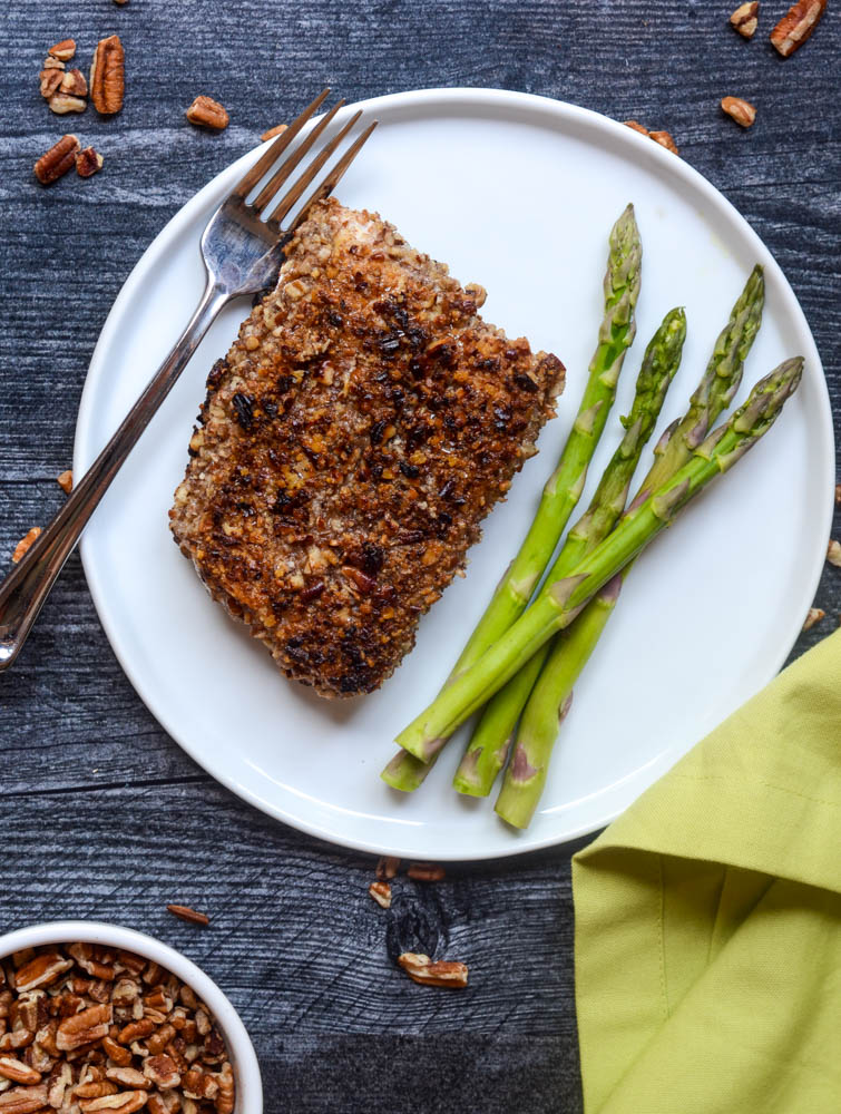 aerial shot of pecan crusted halibut with asparagus on white plate on black wooden background with scattered pecans. 