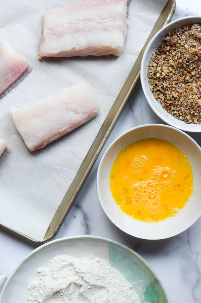 aerial view of naked fish fillets on parchment lined baking sheet, and 3 bowls of flour, egg, and pecan mixture. 