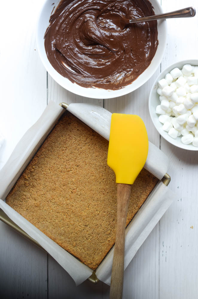 aerial picture of graham cracker crust, bowl of chocolate and marshmallows and yellow spatula. 