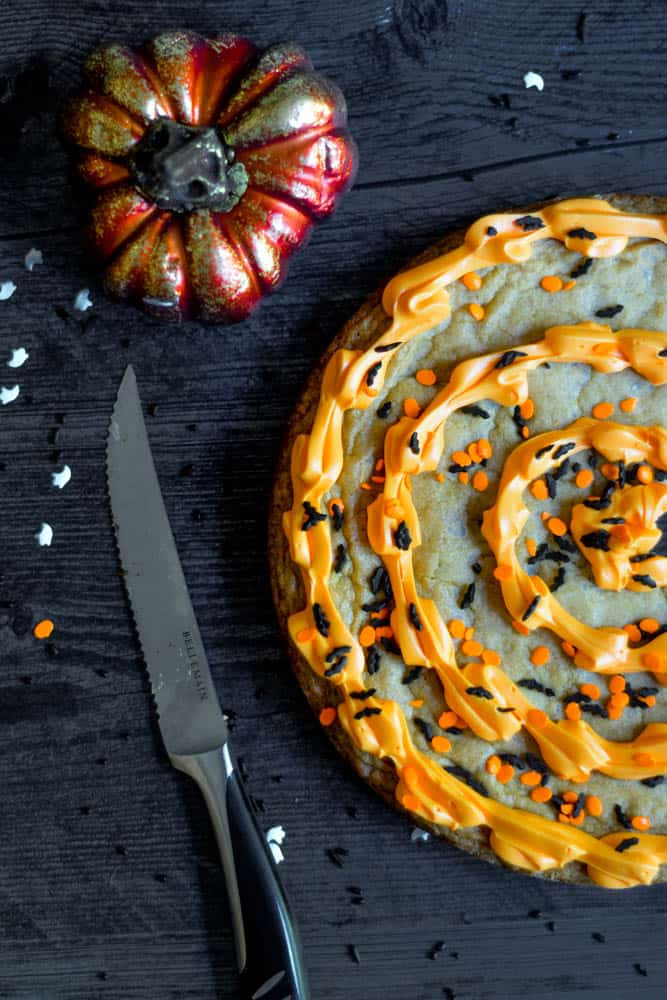 flat lay of cookie cake against black backdrop with knife and pumpkin next to cake.