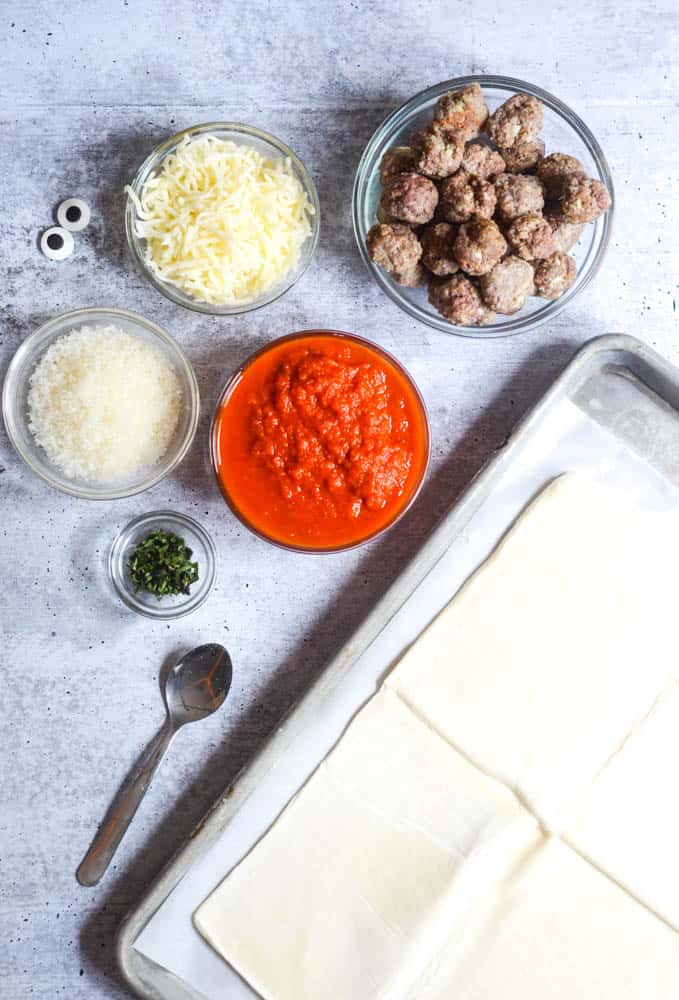 flat lay picture of ingredients needed: puff pastry, basil, marinara sauce, parmesan, mozzarella cheese, meatballs, and candy eyes. 