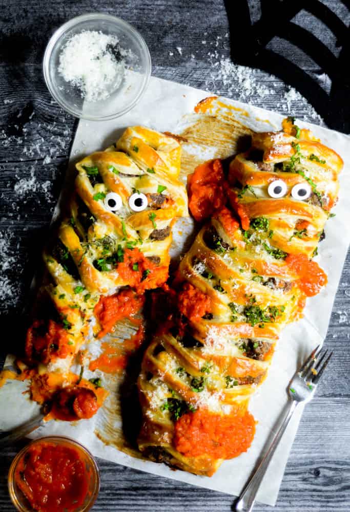 two meatball mummies dressed with marinara, cheese and basil on parchment paper against black wooden background.