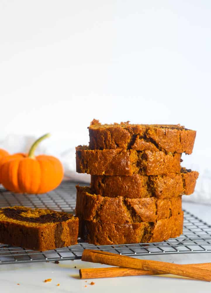 stacked slices of pumpkin chocolate swirl bread on wire rack with pumpkin in background and cinnamon sticks in foreground.