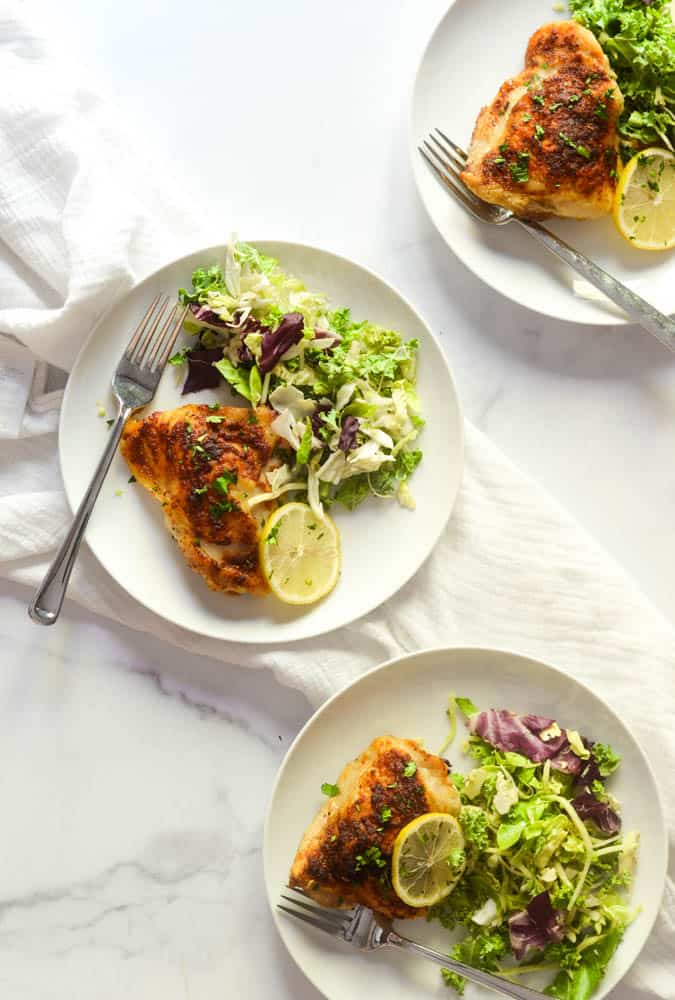 aerial flat flay on three white plates with roasted chicken thigh, side salad, and slice of lemon against marble backdrop. 