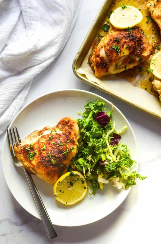 flat lay of one white plate with chicken thigh, side salad, and lemon slice next to sheet pan with other chicken thighs on it. 