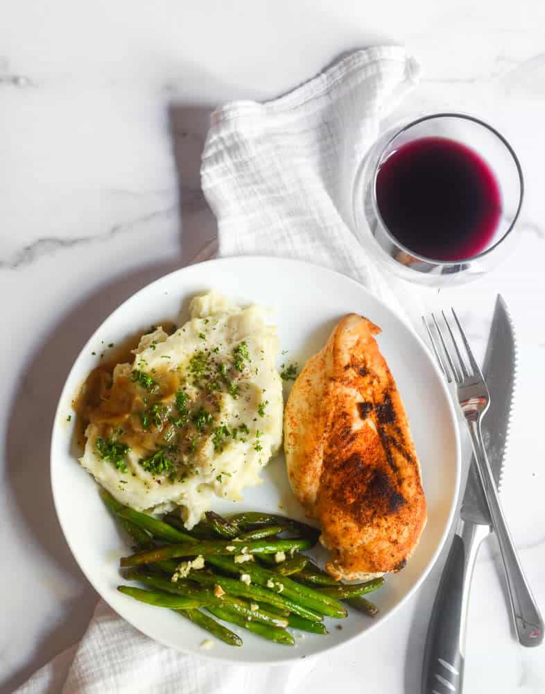 flat lay picture of white plate with dinner on it: chicken breast, mashed potatoes and gravy, and green beans with a glass of red wine. 