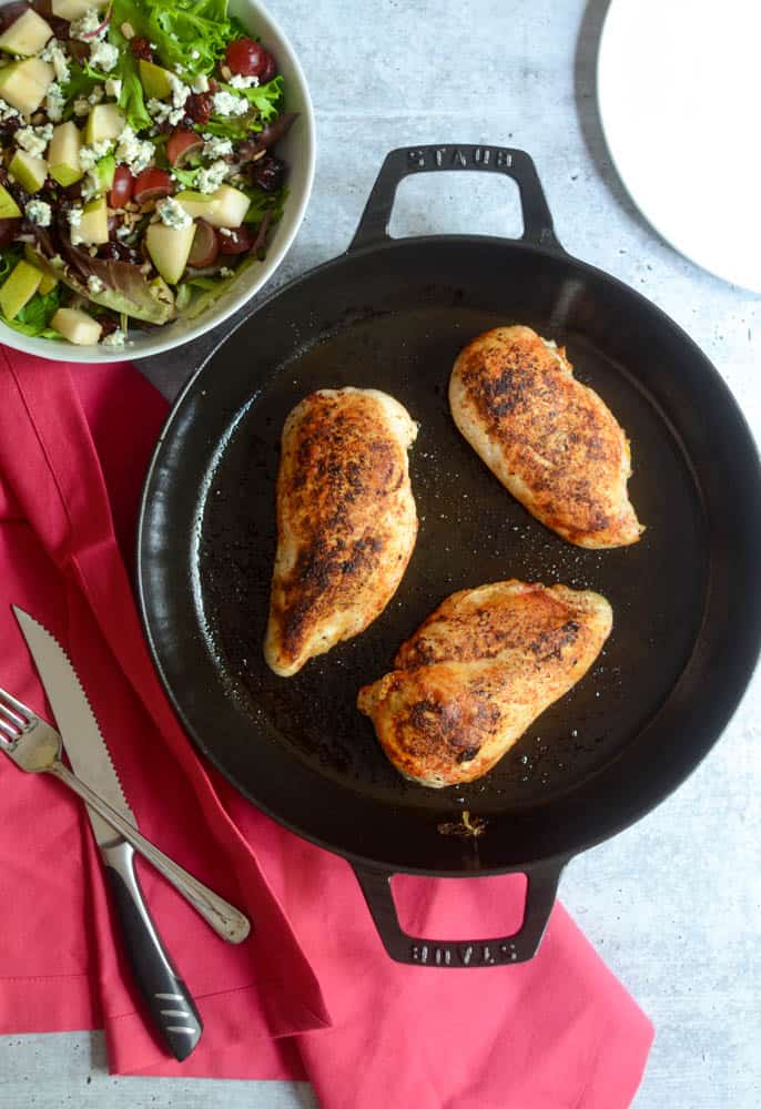 flat lay of 3 seared chicken breasts in cast iron skillet under a pink napkin and side salad in corner.