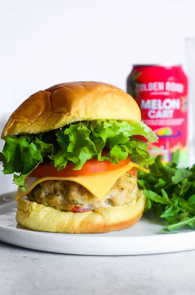 up close picture of baked chicken burger on bun with cheese, lettuce and tomato and a can of beer in the background. 