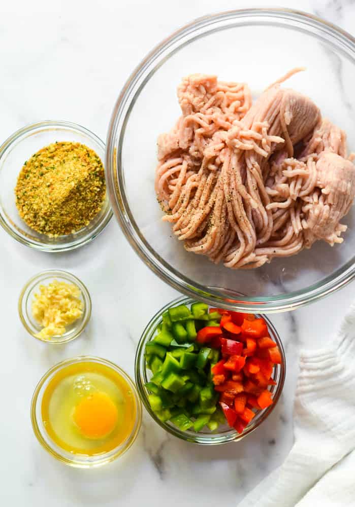 ingredients needed for baked chicken burgers all in separate glass bowls: ground chicken, breadcrumbs, garlic, egg, and bell peppers. 