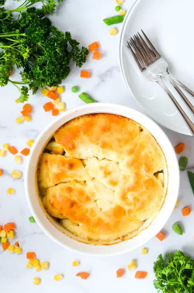 overhead picture of casserole dish of chicken pot pie against a white background with vegetables scattered around. 