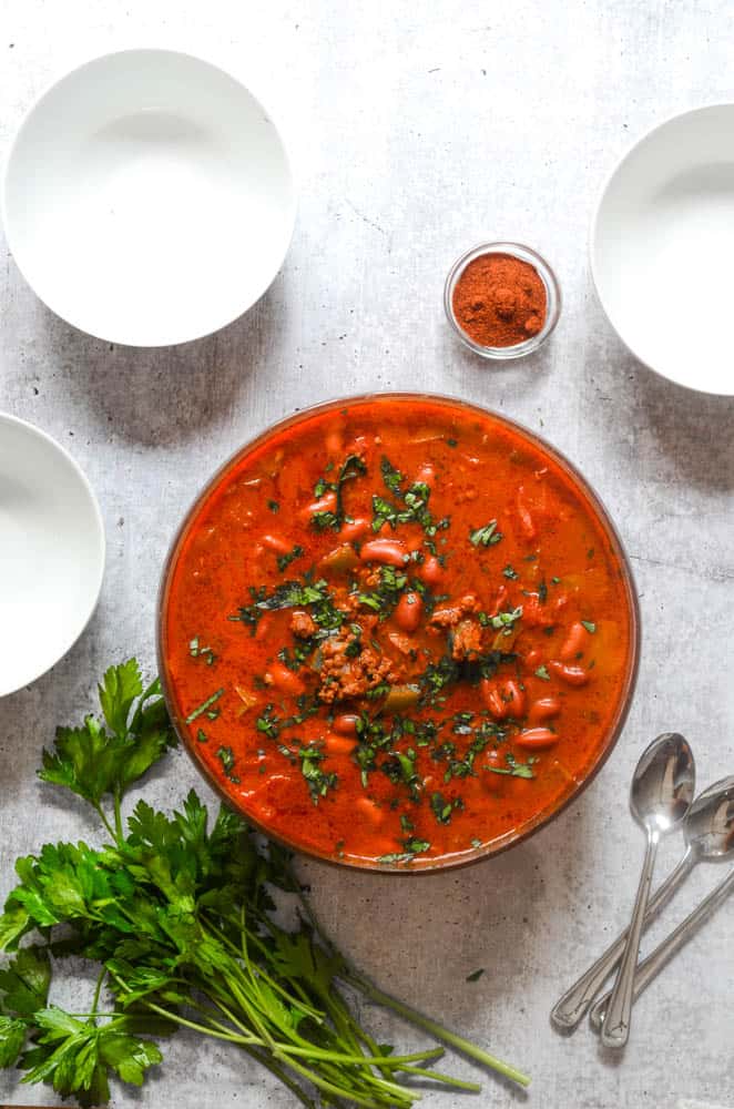 overhead shot of huge bowl of chili with white bowls and bundle of parsley on concrete backdrop. 