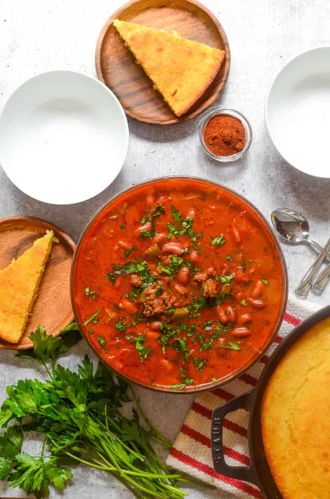 aerial shot of big bowl of chili with empty white bowls and wooden plates with a slice of cornbread on each. 