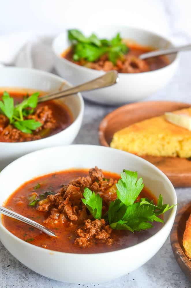 close up picture of one bowl of chili with another bowl of chili and plate of cornbread in the background. 