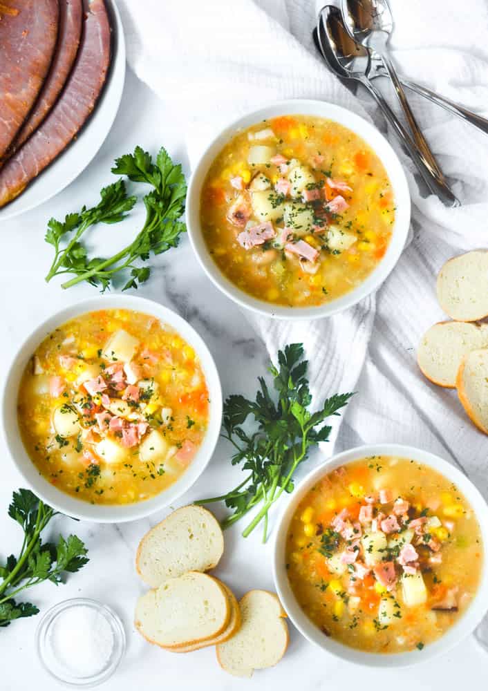 aerial picture of three bowls of ham soup in white bowls against marble backdrop. Parsley and slices of baguette strewn about. 