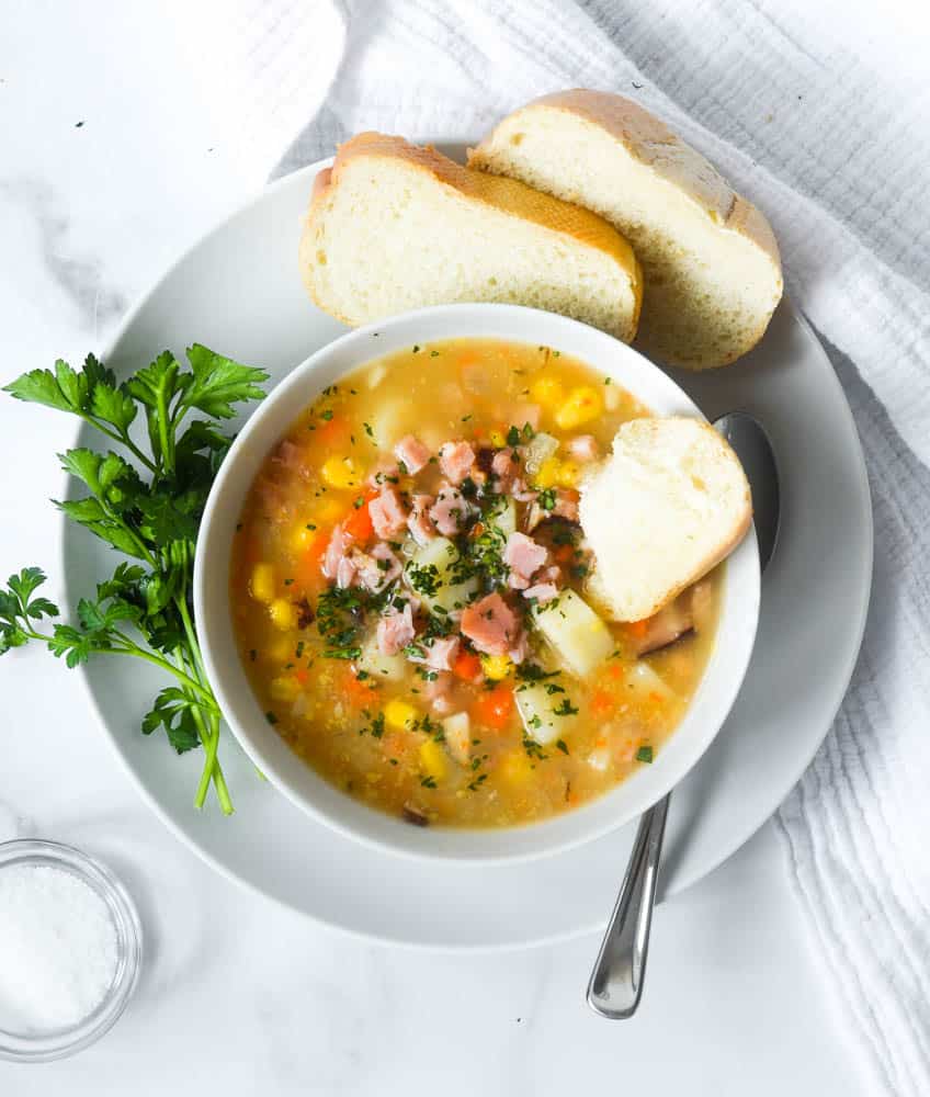 bowl fo ham and potato soup in a white bowl on top of white plate that has parsley sprig and slices of french bread. 