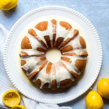 aerial picture of lemon bundt cake on white plate with lemonds around the blue backdrop.