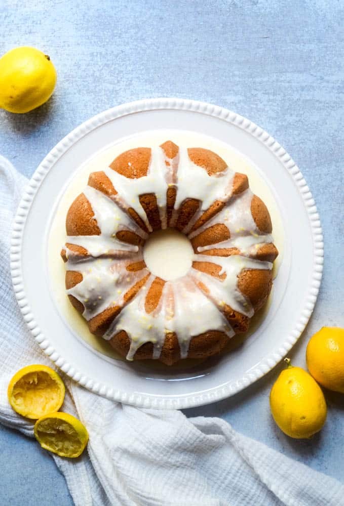 aerial picture of the lemon bundt cake with lemon glaze on circular white platter with white towel and lemons strewn about. 