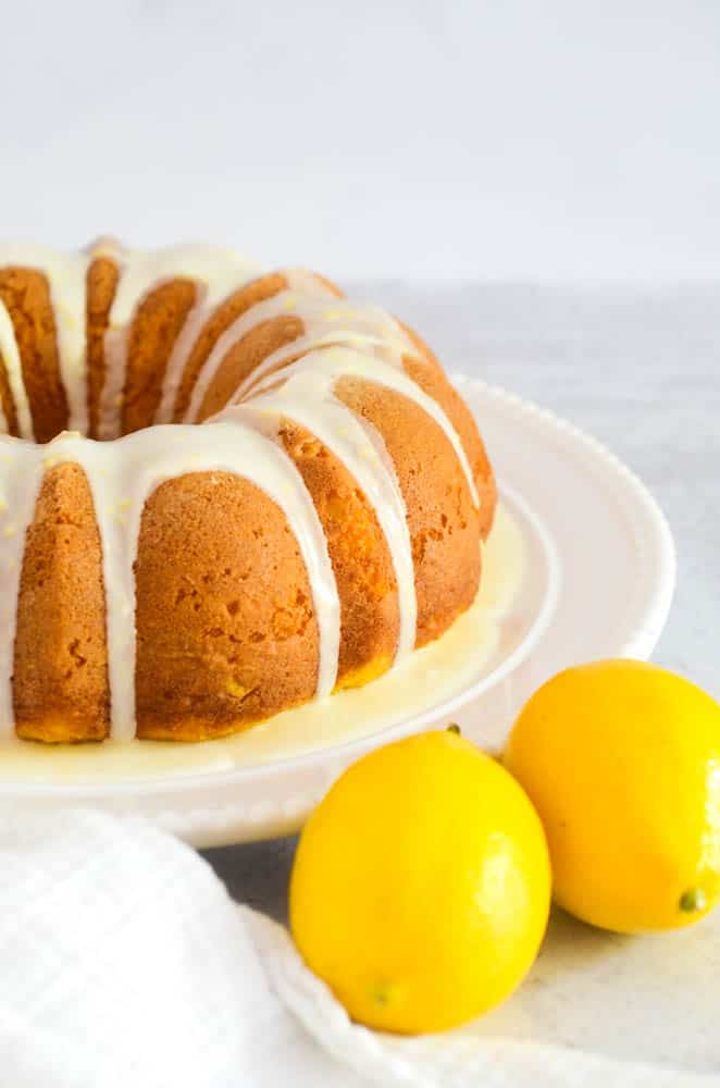 side view of lemon bundt cake on white circular plate with two lemons in the foreground. 