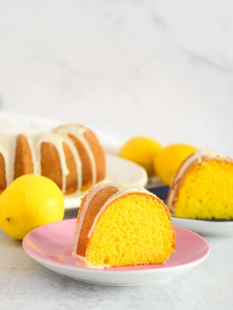 side shot of slice of lemon cake with lemons and another slice of cake in the background. 