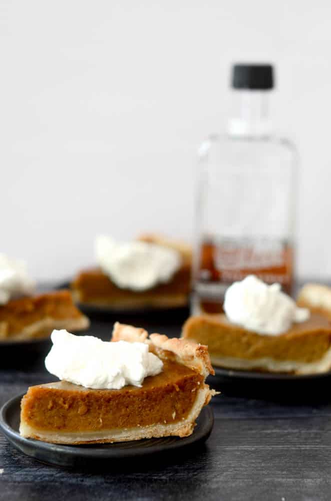 side shot of pumpkin pie slices with bottle of maple syrup blurry in background. 