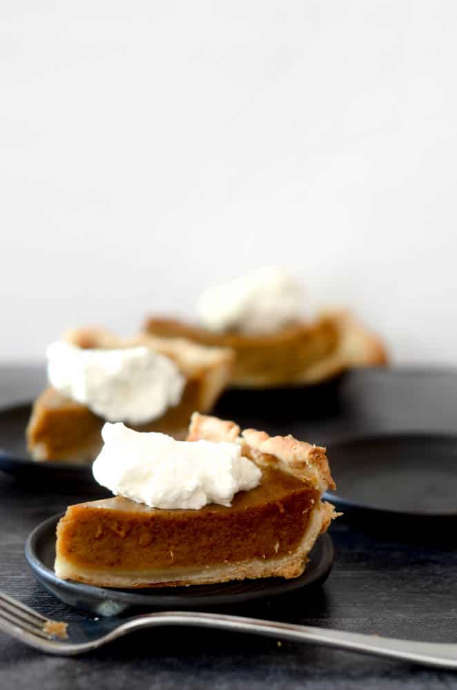 side shot of one slice of pumpkin pie with whipped cream on small black plate. fork is resting in front of plate with other pumpkin pies blurry in background. 