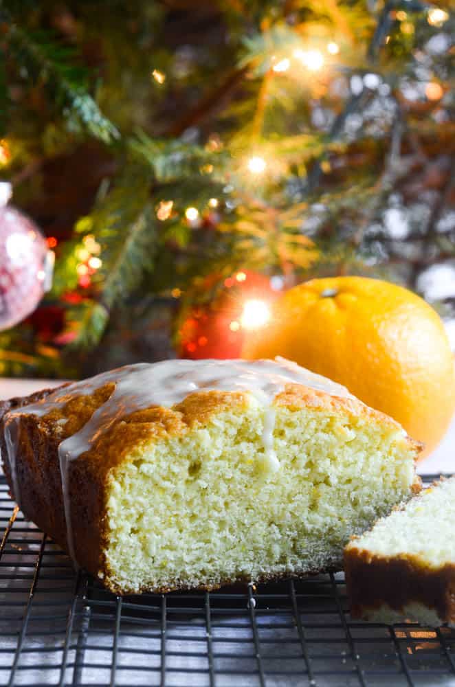 orange pound cake with slice cut into in front of Christmas tree with lights twinkling. 