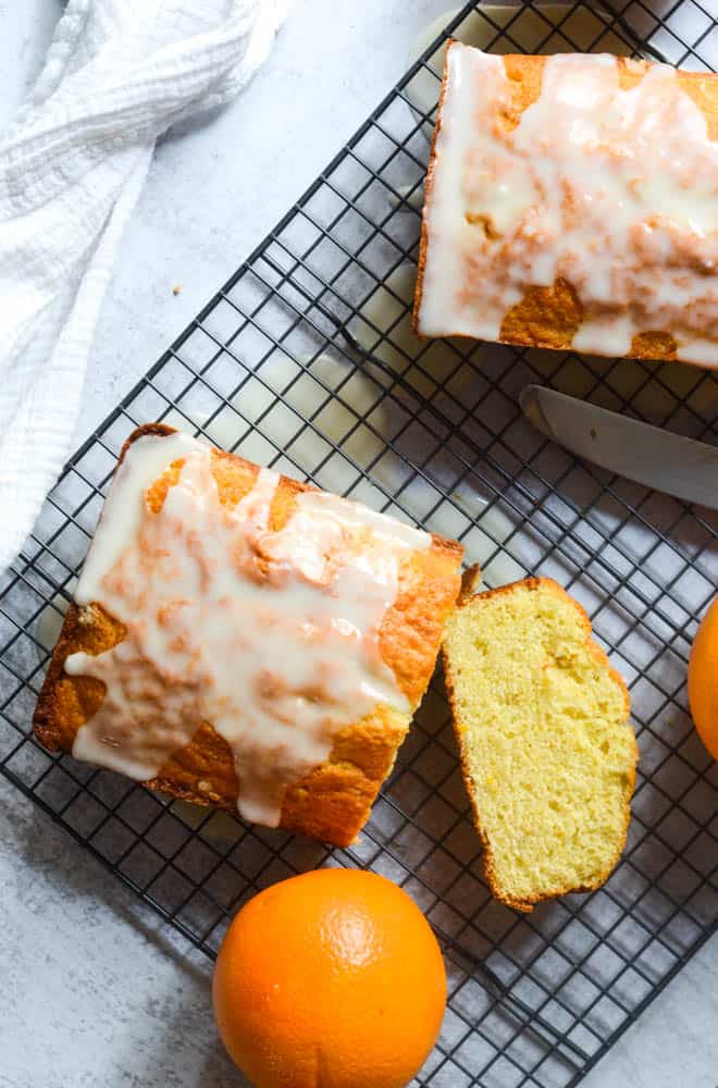 Flat lay of two loaves of orange pound cake on wire rack with one slice cut into and oranges scattered around. 