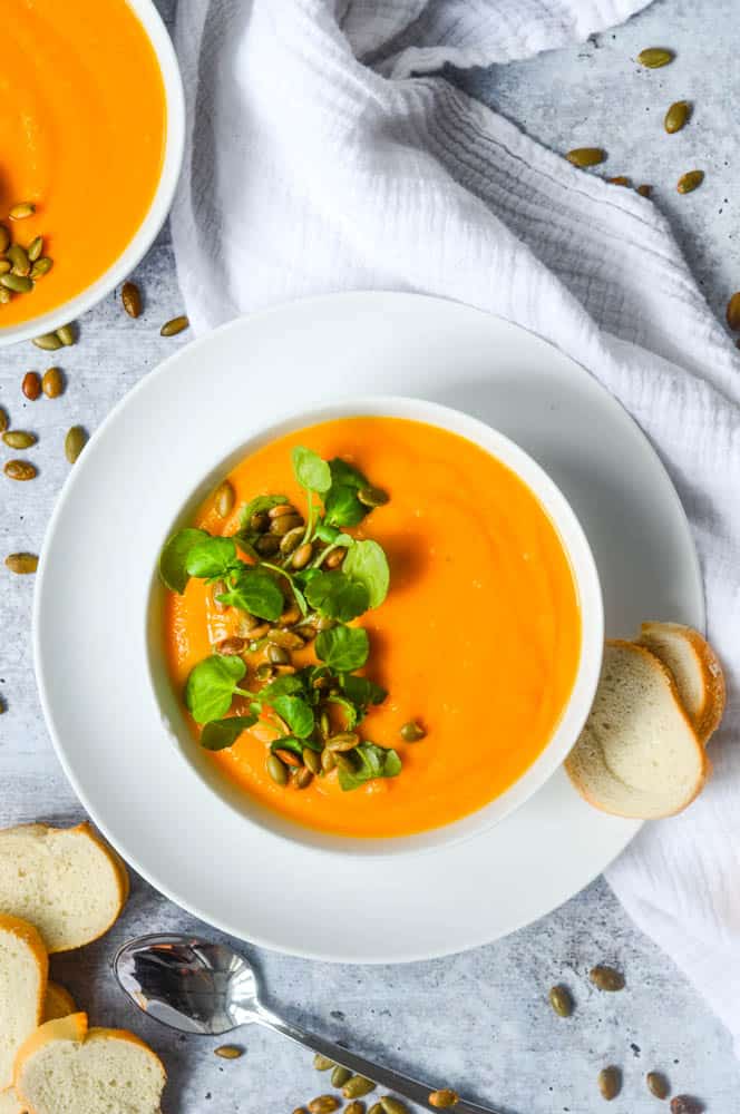 overhead picture of white bowl of soup over one white plate.  Slices of baguette and a spoon are near bow and pepitas are strewn aboutl. 