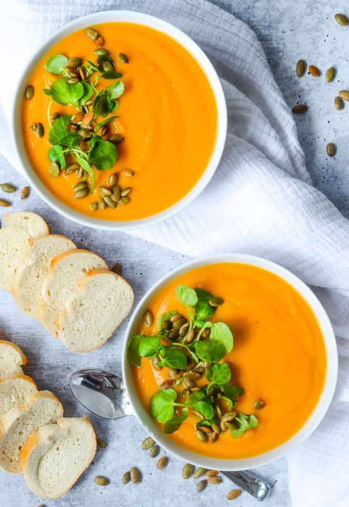 flat lay picture of two large bowls of butternut squash soup in white bowls with slices of baguette strewn around.