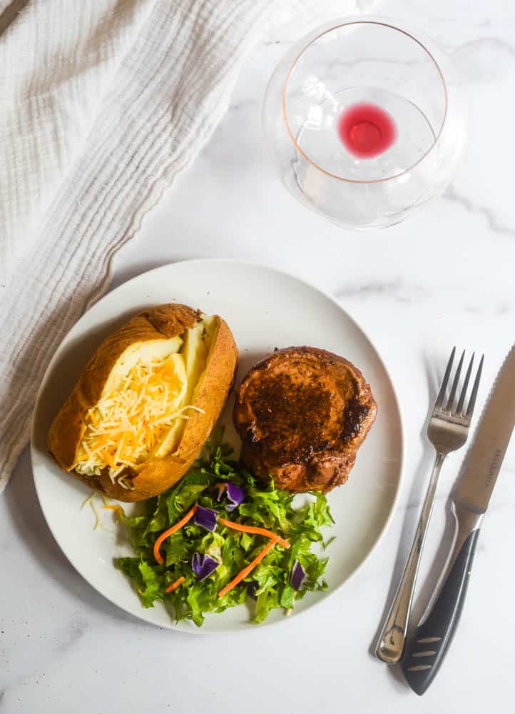 aerial picture of plate of food consisting of filet, salad, and baked potato with fork and knife next to plate, white napkin, and wine glass.