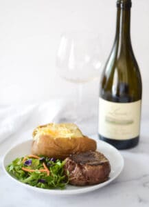 side photo of a plate with filet, baked potato, and salad on it and wine bottle and glass behind plate.