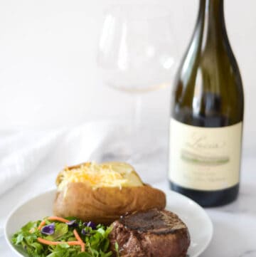 side photo of a plate with filet, baked potato, and salad on it and wine bottle and glass behind plate.