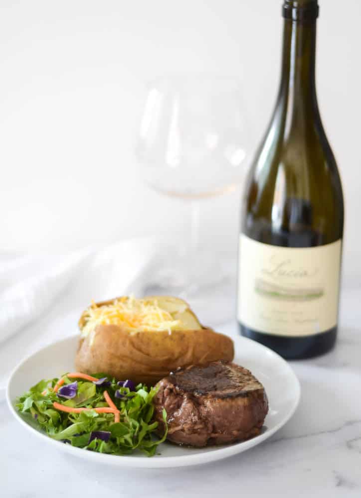 side view of filet on white plate with salad and baked potato. wine bottle and glass in the background.