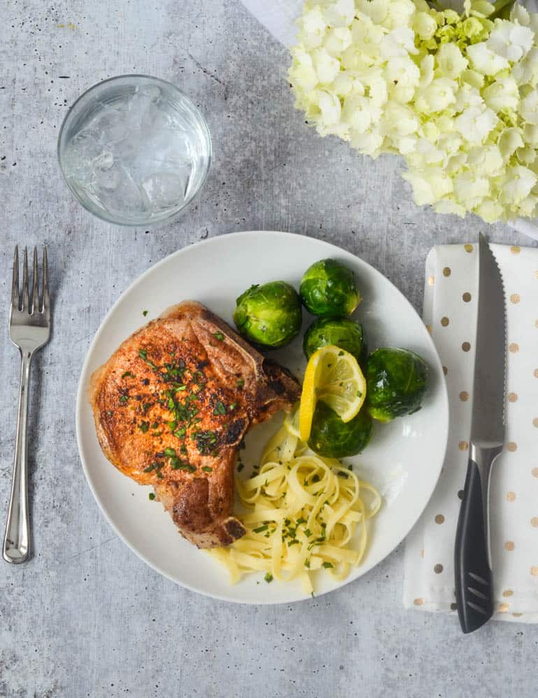 aerial shot of white dinner plate with bone-in pork chop, fettuccine, and brussels sprouts. 