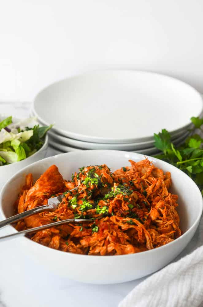 large white bowl of shredded chicken with parsley on top and two forks stuck inside.