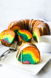 rainbow bundt cake in background with one slice on white square plate