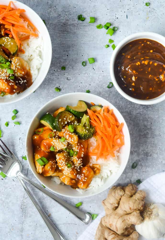 overhead picture of chicken teriyaki bowl with bowl of teriyaki sauce, garlic head, and ginger root next to it. 