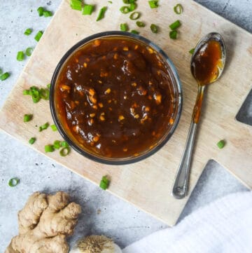 bowl of homemade teriyaki sauce on wooden cutting board surrounded by green onions, garlic, and fresh ginger.