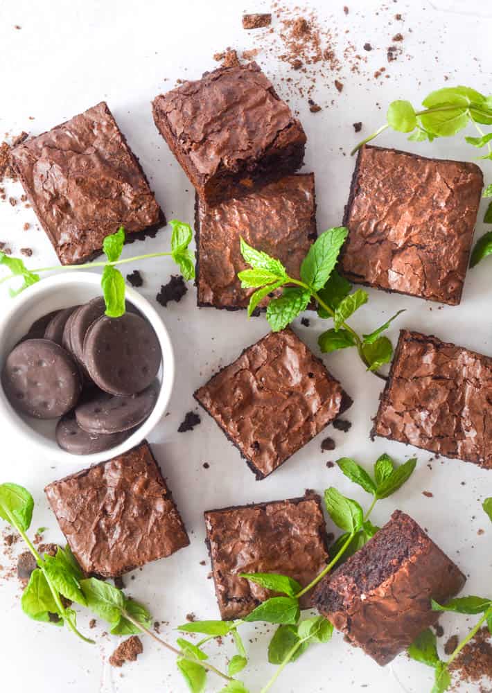 flat lay picture of 9 squares of cut brownies surrounded by mint leaves and a bowl of Thin Mint Girl Scout cookies. 