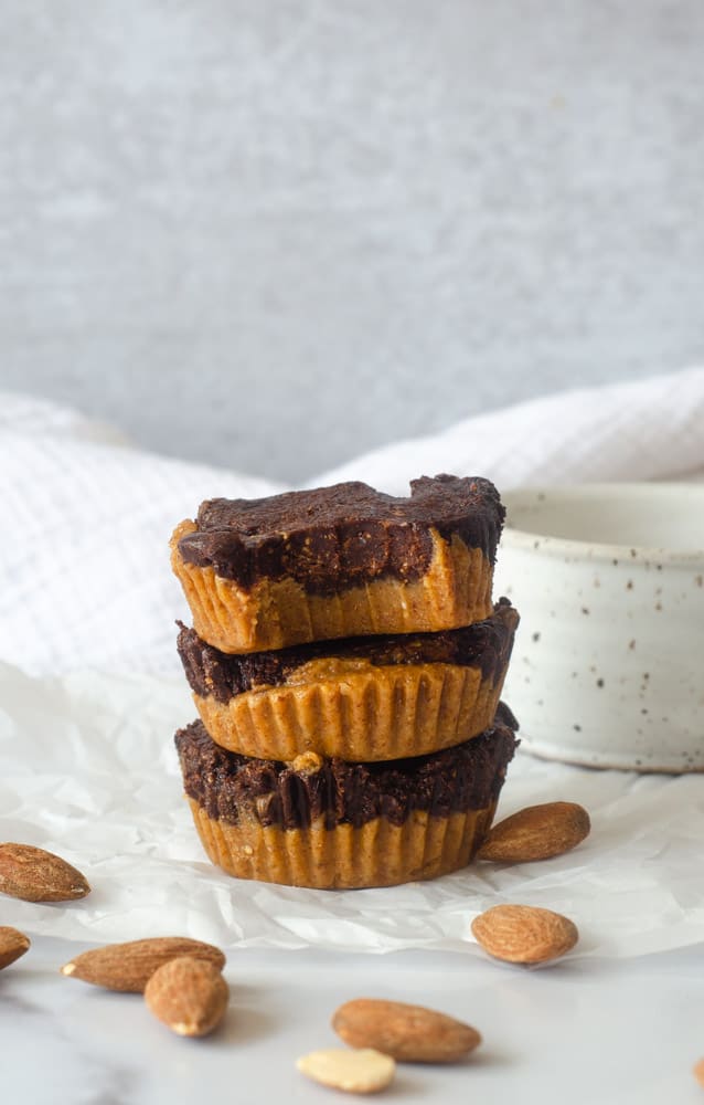 side shot of 3 stacked almond butter cups with the top-most cup bitten into exposing bite marks. Almonds are strewn about. 