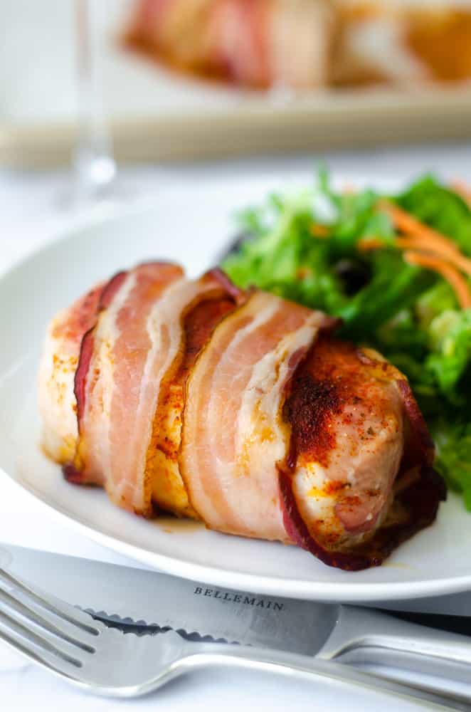 up close picture of bacon wrapped pork chop on white plate with a green salad. 