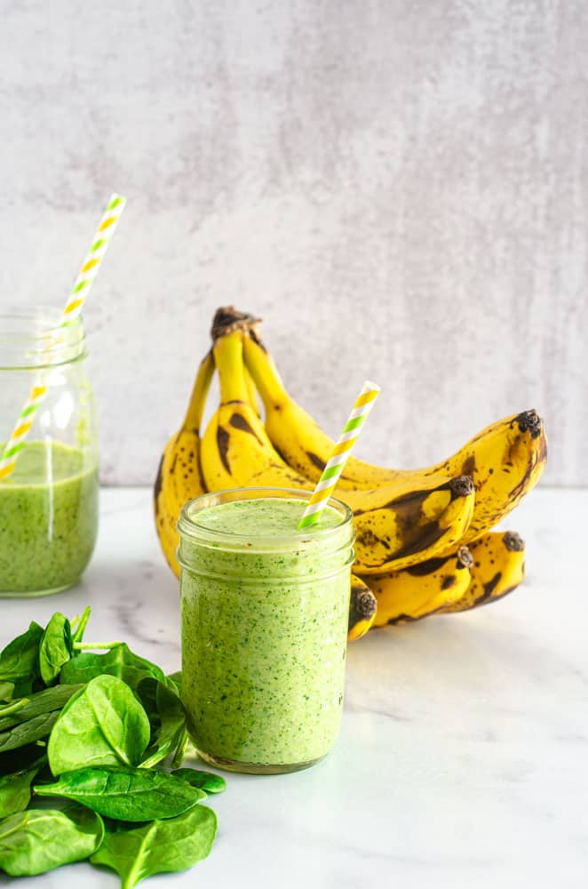 green smoothie with loose spinach next to cup and bananas in the background with another smoothie.