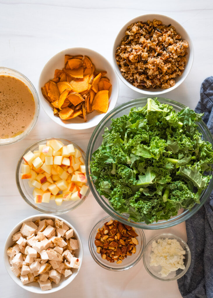 Individual ingredient bowls for the salad, including kale, apples, chicken, almonds, goat cheese, and wild rice.