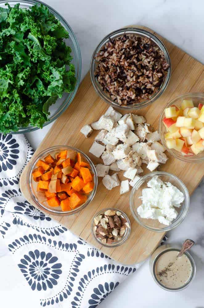 harvest bowl ingredients on a wooden cutting board in glass bowls with large bowl of kale to the side. 