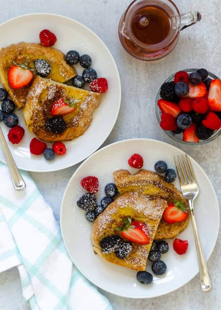 overheat shot of two white plate with french toast, bowl of berries, and maple syrup on light grey background.