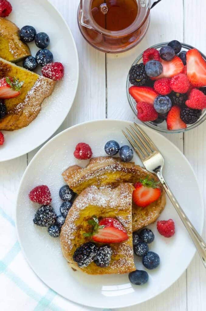 flatlay of two plates of french toast with bowl of fresh berries and carafe or maple syrup. 