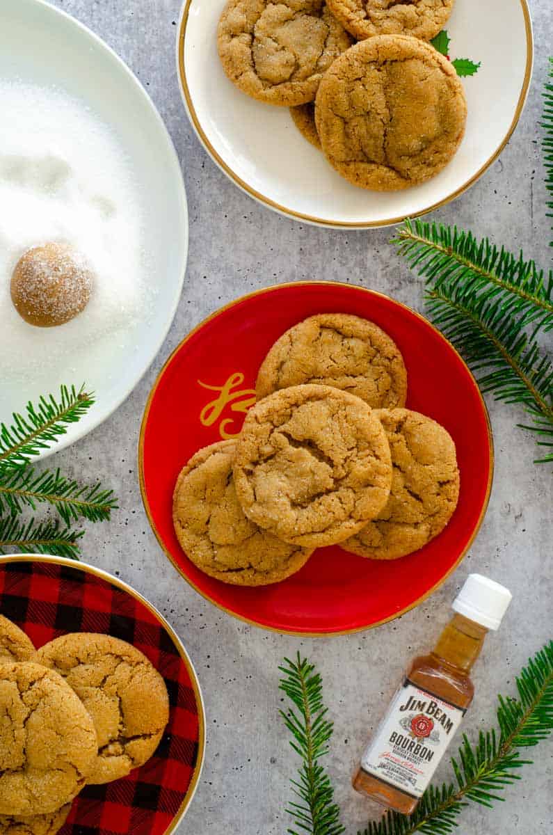 flat lay photo with 3 plates of bourbon ginger molasses cookies next to plate of sugar where dough ball is being rolled