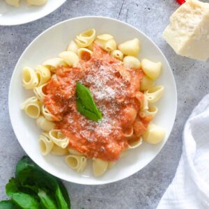 square image of large white plate of rose pasta with parmesan and basil leaf against light blue background.