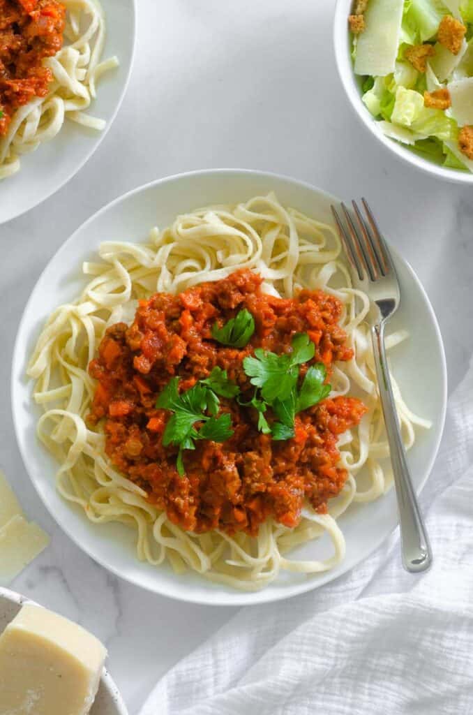 aerial photo of a large plate of pasta with meat sauce on top and parmesan cheese, a caesar salad, and another plate of pasta nearby.